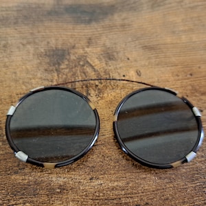May include: A pair of round, dark-tinted clip-on sunglasses. The frames are a dark brown with a tortoiseshell pattern on the outer edges. Silver-colored clips secure the lenses. A thin, dark wire connects the two lenses.