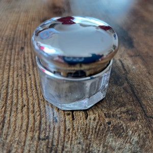 Könnte beinhalten: Ein kleines, klares Glasgefäß mit einem silbernen Deckel. Das Gefäß hat ein strukturiertes, facettiertes Design. Der Deckel ist glänzend und reflektiert das Licht.