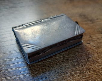 May include: A small, rectangular silver metal box with a hinged lid. The box has a textured surface with etched details and a decorative border. The lid has cursive text. The box is resting on a wooden surface.