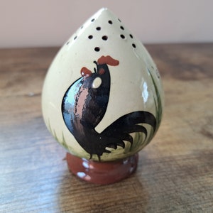 May include: A ceramic rooster salt shaker with a white glaze and a black rooster design. The rooster has a red comb and wattle. The shaker has a hole-punched top for dispensing salt.