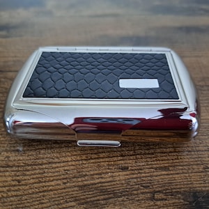 May include: A rectangular metal cigarette case with a polished silver finish and a black textured inlay featuring a repeating scale pattern. The case includes a small, blank, rectangular silver plate on the top surface for personalisation.