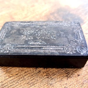 Könnte beinhalten: Eine rechteckige, antike silberne Schatulle mit schwarzem Boden. Der Deckel hat ein verziertes Blumendesign in der Mitte und an den Rändern. Die Schatulle ist wahrscheinlich aus Metall und steht auf einer Holzoberfläche.