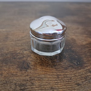 May include: A small, antique glass jar with a silver-colored lid. The jar has a faceted, octagonal shape and a clear body. The lid is domed and reflects the light. The jar is likely for storing small items or cosmetics.