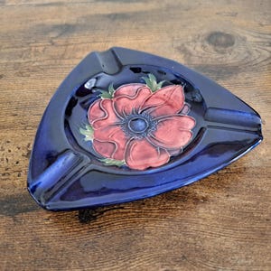 May include: A triangular ceramic ashtray with a glossy dark blue glaze. The centre features a hand-painted red flower with green leaves and a dark blue circular core. The piece has three distinct notches along the edges for resting items.
