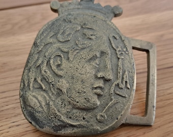 May include: A vintage brass belt buckle featuring a detailed profile of a person wearing a crown. The buckle has a rectangular loop and is set against a wooden surface. The buckle's design is an oval shape with a crown on top.