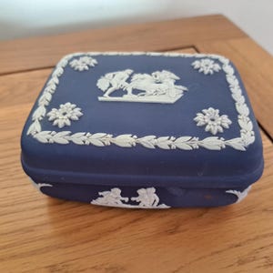 Op de afbeelding: Een marineblauwe rechthoekige sieradendoos met witte reliëfdetails. De doos heeft een decoratieve rand van bladeren en bloemen, met een centraal reliëf van figuren en een strijdwagen. Extra reliëf figuren staan op de basis.