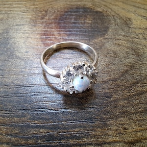 Könnte beinhalten: Ein silberner Ring mit einem weißen Opal-Edelstein, der von kleineren weißen Edelsteinen umgeben ist.