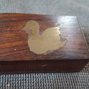 May include: A small wooden box with a brass swan design on the lid. The box is dark brown with a wood grain pattern.