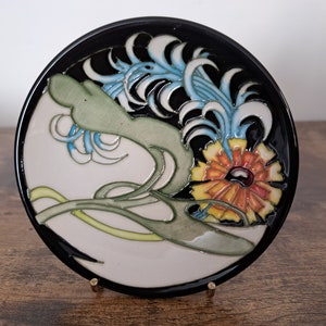 May include: A round ceramic plate with a black rim and a white center. The plate features a stylized floral design with blue, green, yellow, and orange colors.