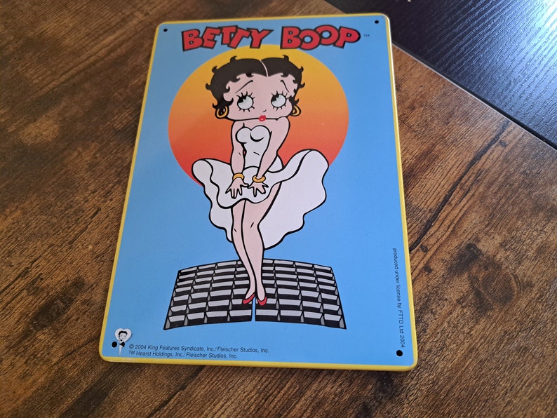 May include: A blue and yellow metal sign featuring a cartoon image of Betty Boop wearing a white dress and standing on a black and white checkered platform. The sign includes the text "BETTY BOOP" and copyright information.