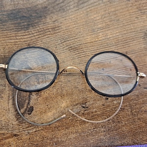 May include: Round eyeglasses with black rims and gold-colored accents. The lenses are clear, and the frame has a delicate, vintage aesthetic. The glasses are resting on a wooden surface.