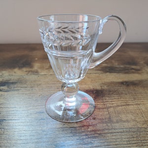 May include: A clear glass cup with a handle and a decorative etched design. The cup has a round base and a stem.