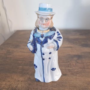 以下が含まれることがあります： 水色の帯が付いた白いシルクハット、丸いワイヤーフレームの眼鏡、濃い青色の花柄のアクセントとボタンと襟が付いた長い白いコートを身に着け、片手に濃い青色のボトルを持っている人物の陶器製の置物。