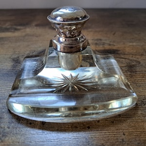 Op de afbeelding: Een inktpot van helder glas met een zilverkleurig deksel. De inktpot heeft een stervormig ontwerp dat in het glas is geëtst.