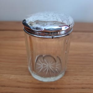 以下が含まれることがあります： 銀色の蓋が付いた透明なガラス瓶。瓶の底にはスターバースト模様があります。蓋はハンマー加工された質感と銀色の縁が付いた銀色です。瓶は木製の表面に置かれています。