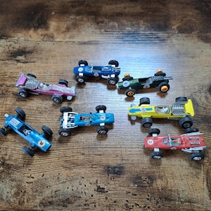May include: A collection of vintage toy race cars in various colors, including blue, pink, green, yellow, and red. Each car has a unique number and design, with visible engine details. The cars are arranged on a wooden surface.