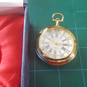 Puede incluir: Un reloj de bolsillo dorado con esfera blanca y números romanos negros. El reloj tiene un movimiento de cuarzo y se presenta en una caja azul con forro de satén rojo. El reloj está sobre una superficie verde.