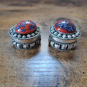 Könnte beinhalten: Zwei kleine, runde, silberfarbene Dosen mit rot-blauen Mustern auf den Deckeln. Die Dosen haben aufwendige Details an den Seiten und Oberseiten und stehen auf einer Holzoberfläche. Sie scheinen dekorativ zu sein oder zur Aufbewahrung kleiner Gegenstände.