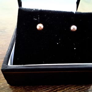 Puede incluir: Un par de pendientes de botón pequeños y redondos presentados en una caja de joyería negra con un interior de terciopelo negro. Los pendientes son de color oro claro y contrastan con el fondo oscuro de la caja.