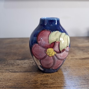 May include: A small, blue ceramic vase with a floral design. The vase features a large, stylized flower in shades of pink, yellow, and green. The vase is approximately 15 cm tall and has a glossy finish.