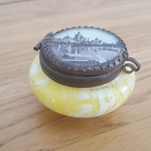 以下が含まれることがあります： 金属製の蓋が付いた、黄色と白の小さなガラス製の宝箱。蓋には、橋のある都市の白黒写真が貼られています。