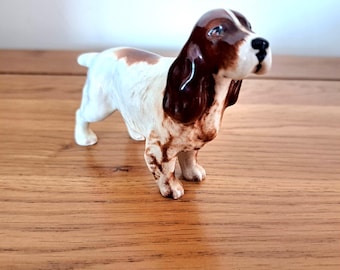 May include: A ceramic dog figurine in a standing pose. The dog has a white body with brown patches on its head and back. The ears and top of the head are a dark brown. The dog's nose is black, and it has a small tail.
