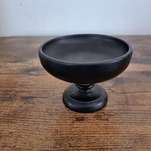 May include: A small, matte black pedestal bowl with a smooth, rounded basin and a sturdy, circular base. The object features a uniform dark finish throughout, with a simple, clean silhouette and a wide, open top for holding small items.