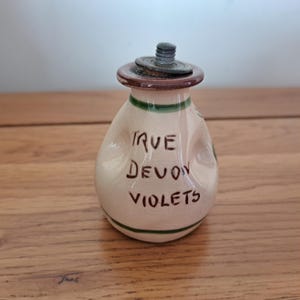 May include: A small, off-white ceramic bottle with a brown lid and green accents. The bottle has the words "TRUE DEVON VIOLETS" written on it in brown script. The bottle is sitting on a wooden surface.