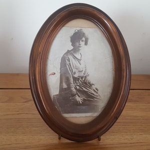 May include: An oval wooden frame with a black and white photograph of a woman in a dress. The frame is on a wooden surface.