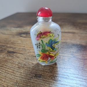 Può includere: Una piccola bottiglia di vetro smerigliato con un tappo rosso. La bottiglia è decorata con tre pesci rossi colorati e caratteri cinesi.