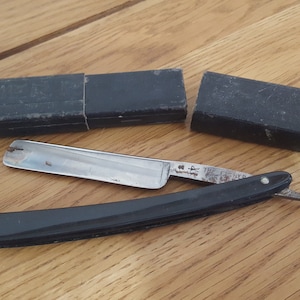 May include: A vintage straight razor with a black handle and a silver blade. The blade is inscribed with "The Fischer" and a logo. The razor is in a black leather case.
