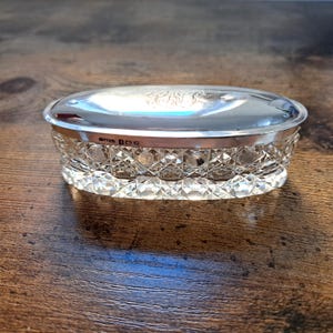 Peut inclure: Une boîte à bijoux ovale en cristal et argent. La base en cristal transparent présente un motif taillé en diamant, et le couvercle est en argent poli avec des initiales gravées. La boîte est posée sur une surface en bois.