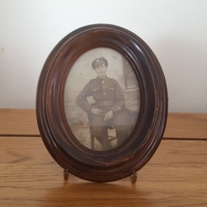 May include: A black and white photo of a man in a military uniform, wearing a cap, sitting on a chair. The photo is framed in a dark wood oval frame.