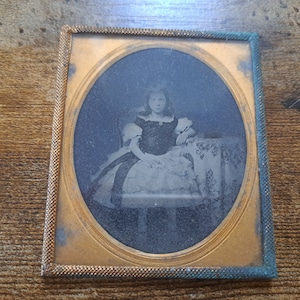 Op de afbeelding: Een zwart-wit portret van een jong meisje dat een jurk draagt en op een stoel zit. Het portret is ingelijst in een gouden lijst met een blauwe rand.