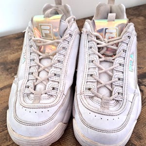 以下が含まれることがあります： 白のFila Disruptor IIスニーカーのペア。分厚いソール、レースアップ開閉、タンのイリデセント素材のFilaロゴが特徴です。スニーカーは木製の表面にあります。
