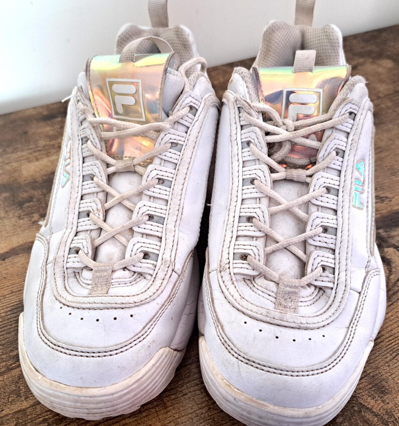 以下が含まれることがあります： 白のFila Disruptor IIスニーカーのペア。分厚いソール、レースアップ開閉、タンのイリデセント素材のFilaロゴが特徴です。スニーカーは木製の表面にあります。