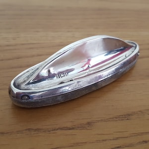 Peut inclure: Un objet décoratif en argent en forme de bateau avec une surface lisse et réfléchissante. L'objet a un petit détail rond et surélevé à chaque extrémité. L'objet est marqué du texte "STERLING 925".