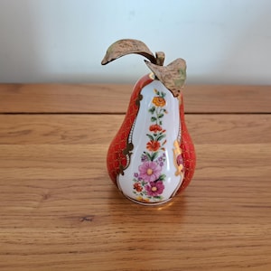May include: A decorative pear figurine with a red lattice pattern and a white floral design. The pear has a gold stem and leaves. The figurine is made of ceramic and is approximately 6 inches tall.