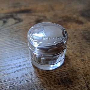 Könnte beinhalten: Ein kleiner, runder, versilberter Behälter mit Glasboden. Der Deckel hat einen dekorativen Rand und die Inschrift "STERLING" ist auf der Oberseite sichtbar.