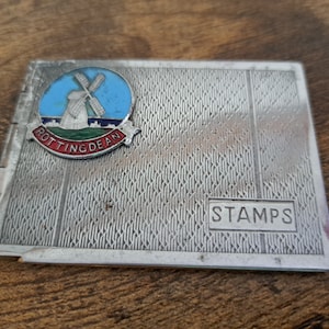 Peut inclure: Un album de timbres en métal argenté avec une épingle en émail représentant un moulin à vent de Rottingdean sur le devant. L'album a une surface texturée et le mot "STAMPS" est imprimé sur le devant.