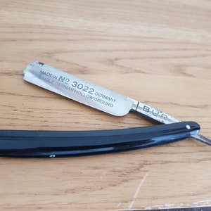 Może przedstawiać: Brzytwa z czarnym uchwytem i srebrnym ostrzem. Ostrze jest grawerowane napisem "Made in Germany No. 3022 Single German Hollow Ground" i "Bur".