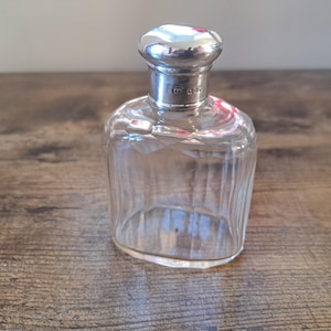 Könnte beinhalten: Eine klare Glasflasche mit einem silbernen Verschluss. Die Flasche hat eine strukturierte Oberfläche und der Verschluss trägt eine Punze.