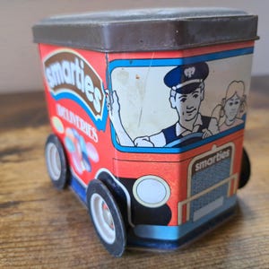 Könnte beinhalten: Eine vintage Blechdose in Form eines Lieferwagens mit dem Schriftzug "Smarties Deliveries" an der Seite. Der LKW ist rot und blau und hat einen Fahrer mit blauer Mütze und ein Mädchen auf dem Beifahrersitz. Die Dose ist aus Metall und hat einen braunen Deckel.