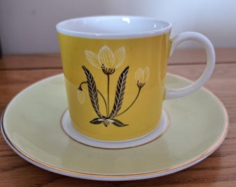 May include: A yellow teacup and saucer set. The cup features a floral design in black and white. The saucer and the inside of the cup are white. The set has a gold trim.