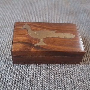 Può includere: Una piccola scatola di legno con un'intarsio in ottone di un uccello. La scatola ha una venatura del legno marrone scuro.