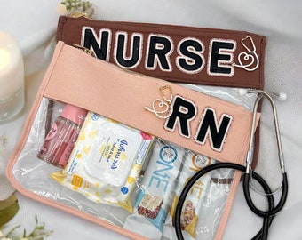 May include: A clear plastic zippered pouch with a pink fabric lining. The pouch has a brown fabric flap with the word "NURSE" in black letters. The pouch contains a stethoscope, hand sanitizer, wipes, and snacks.