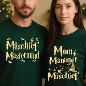 May include: Two people wearing matching dark green sweatshirts. The sweatshirt on the left says "Mischief Mastermind" in gold lettering. The sweatshirt on the right says "Mom Manager of Mischief" in gold lettering, with stars and a wizard hat.
