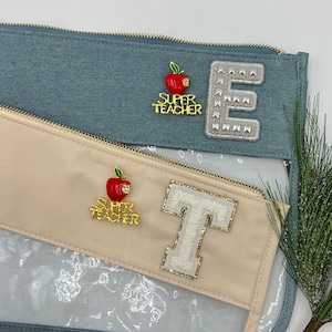 May include: Two clear zippered pouches with a light blue and beige fabric exterior. The pouches have a clear plastic window on the front. The blue pouch has a gold "E" with white rhinestones and a red apple pin with the words "Super Teacher". The beige pouch has a gold "T" with white rhinestones and a red apple pin with the words "Super Teacher".