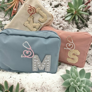 Puede incluir: Tres bolsas de tela con cremallera con diferentes letras y adornos. Las bolsas son azules, rosas y beige. La bolsa azul tiene un estetoscopio plateado con un corazón rosa y una letra M plateada con pedrería. La bolsa rosa tiene un estetoscopio plateado con un corazón blanco y una letra S dorada. La bolsa beige tiene un corazón rojo y una letra K y J dorada.