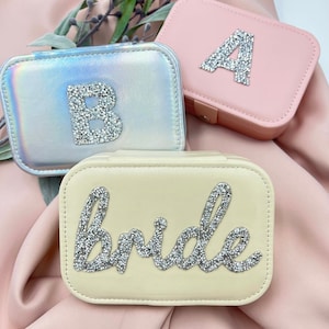 Könnte beinhalten: Drei kleine Schmuckkästchen in Pastellfarben. Eines ist schillernd mit einem kristallbesetzten "B", ein anderes ist rosa mit einem Kristall-"A", und das dritte ist cremefarben mit dem Wort "bride" in Kristallen. Die Kästchen sind rechteckig mit abgerundeten Ecken.